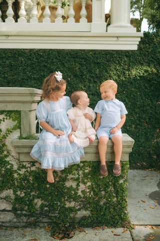 Three children wearing Little Harts clothing, a big sister in a smocked dress, baby in a dress, and toddler boy in a classic set - showcasing quality, Southern-inspired styles from the New Arrivals collection.