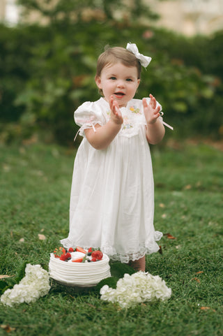 Toddler girl wearing a white heirloom-style Celebration Dress with lace detailing, hand embroidery, and a scalloped hem, perfect for special occasions.