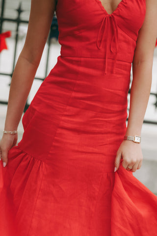 Woman wearing a sleeveless linen midi dress with gathered straps, tie-front neckline, and soft pleating, styled for a timeless and polished look.