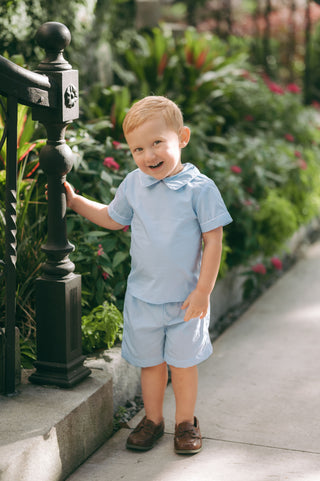 Little boy in light blue piped Peter Short Set with Peter Pan collar, standing in a garden setting — perfect for Easter or special occasion dressing.