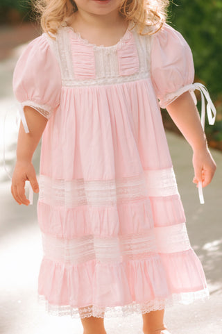 Little girl in a light pink Savannah Heirloom Dress from Little Harts, featuring lace, details, puff sleeves with satin bows, and tiered ruffle skirt.