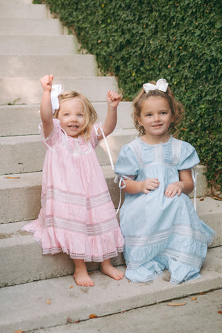 Two little girls wearing the Savannah Heirloom Dress from Little Harts in pale blue and light pink, featuring lace details and puff sleeves with satin bows.